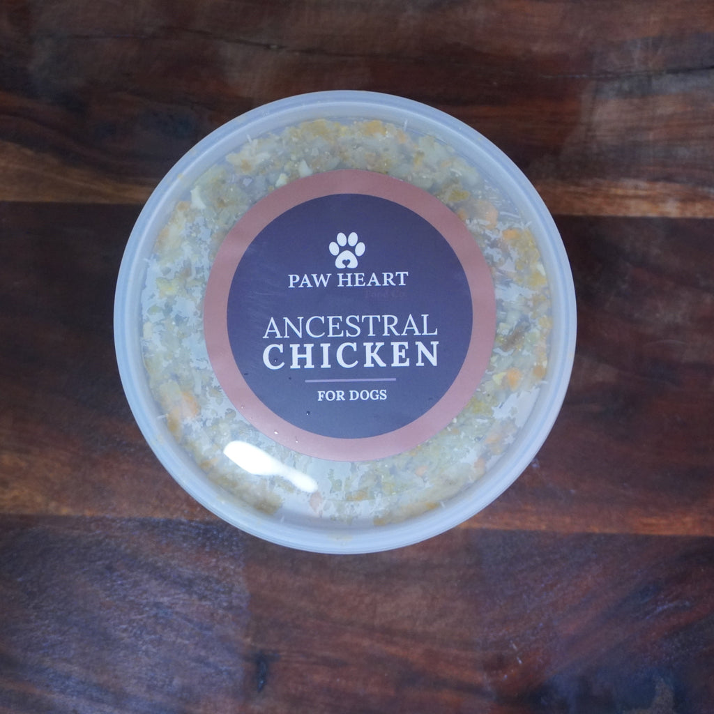 A round plastic tub of Paw Heart Ancestral Chicken wet dog food, featuring a dark brown label with a paw print logo, placed on a dark wooden surface.